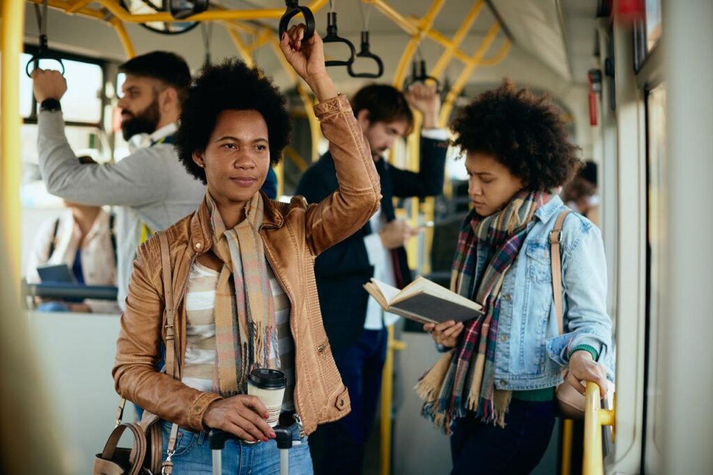 num carro de metrô pessoas estão se segurando. uma mulher lê encostada à parede